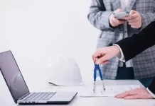 Construction professionals reviewing blueprints with a compass, laptop, and safety helmet on the desk.