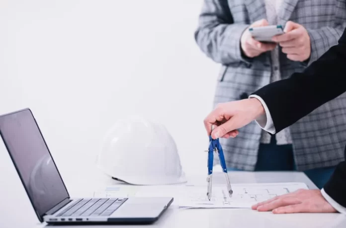 Construction professionals reviewing blueprints with a compass, laptop, and safety helmet on the desk.